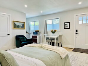Un dormitorio con cama, escritorio y silla. Hay una ventana con cortinas, un cuadro en la pared y una puerta.