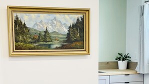 A room with a green velvet armchair, a painting of a mountain landscape, a white cabinet, and a potted plant.