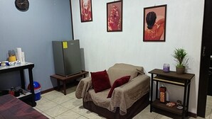 Ein kleiner, möblierter Raum mit einer Couch, einem Tisch, einem Kühlschrank und Wandbildern.
