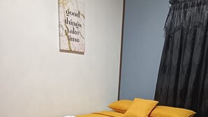 Ein ordentlich bezogenes Bett mit einer gelben Tagesdecke und zwei Kissen, ein weißes Handtuch liegt gefaltet darauf, und ein Schild an der Wand zeigt die Aufschrift 'good things take time'.