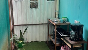 Ein kleiner, abgegrenzter Außenbereich mit einer Wellblechwand, einem grünen Rasenteppichboden, einem Holztisch mit Kaffeemaschine, einem Laptop und einer roten Mülltonne.