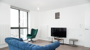Ein modernes Wohnzimmer mit einem blauen Sofa, einem grünen Sessel, einem Flachbildfernseher und einem kleinen Hocker.