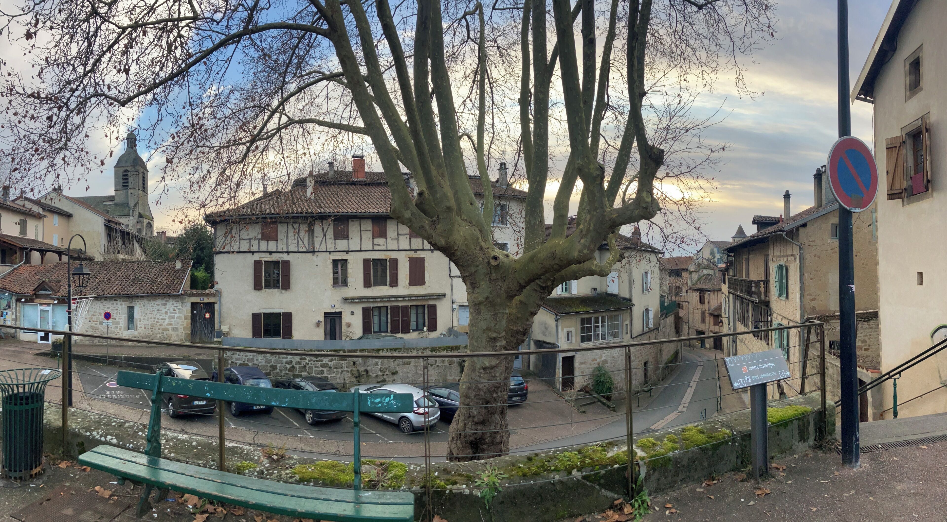 Une rue bordée d’arbres, avec des bâtiments historiques, un panneau indiquant qu’il est interdit de se garer et un banc.