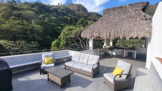An outdoor seating area with wicker furniture and cushions, a thatched-roof structure, and a view of a lush green hillside.