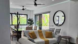 A modern living room with a sofa, coffee table, and chairs. There is a large window with a view of greenery outside.