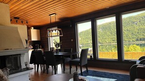 A cozy dining area with a table and chairs, a fireplace, and large windows offering a view of a forested landscape. - Nesset Fiskfjord. House by the sea with panoramic views, Gateway to Lofoten! (Troms)