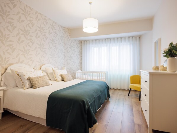 A bedroom with a bed, a crib, a yellow chair, a dresser, and a window with curtains. - Piso 2 Adeinés Viviendas Uso Turístico medio (A Estrada)
