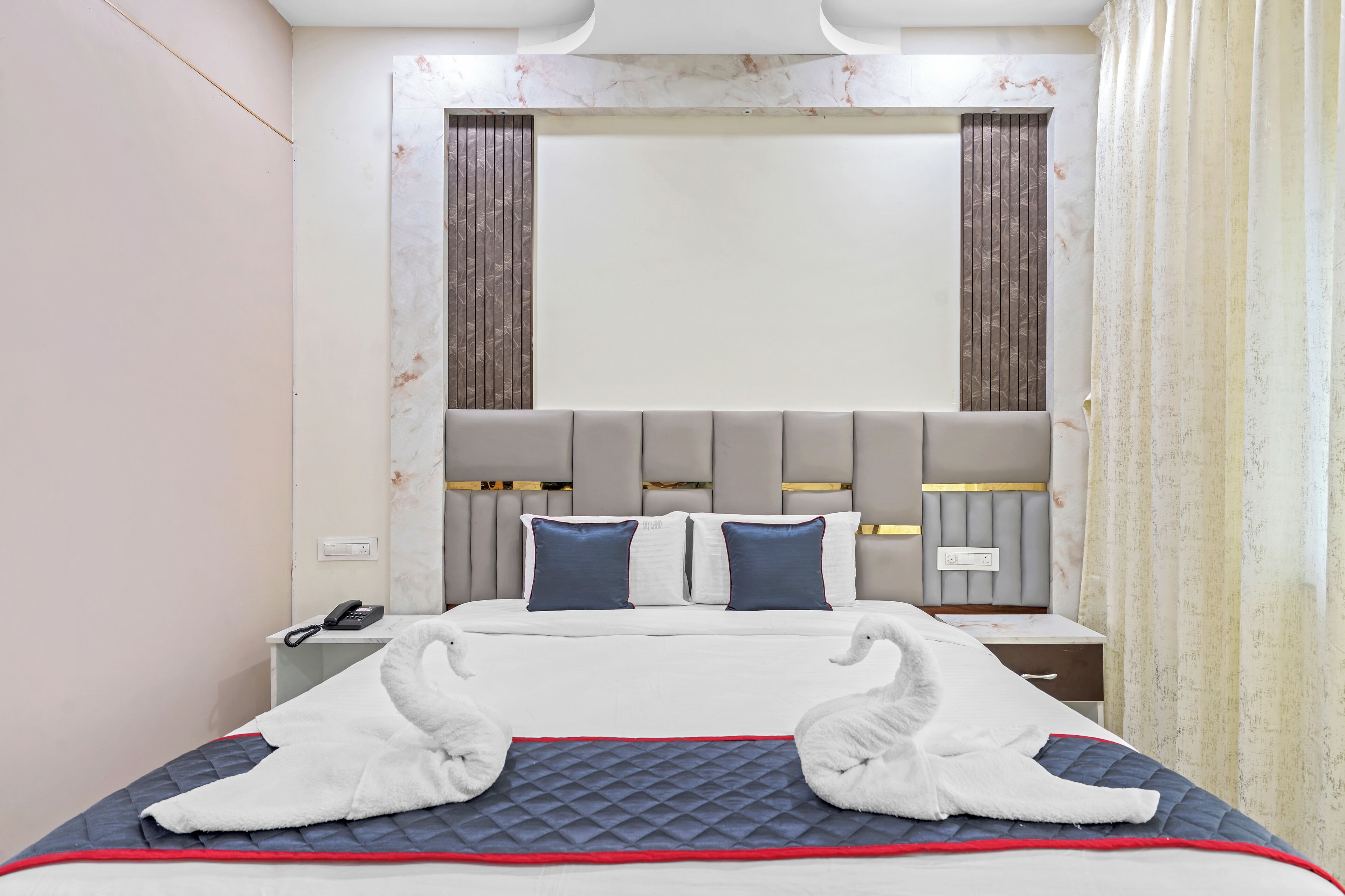 A hotel room with a large bed, a headboard with vertical panels, a bedside table with a phone, and a wall-mounted light fixture.