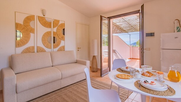Un salón moderno con un sofá color beige, una mesa de comedor dispuesta para el desayuno y vistas al exterior a través de una puerta abierta.