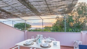 Un área de comedor al aire libre con una mesa preparada para cuatro personas, rodeada de sillas blancas, bajo una pérgola con vistas a árboles y un atardecer.