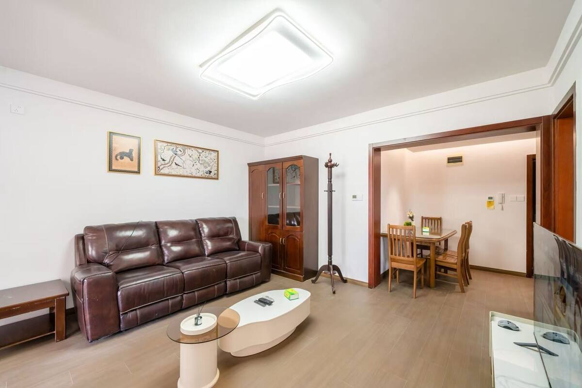 A living room with a brown leather sofa, a white coffee table, a wooden cabinet, and a dining area with a table and chairs.