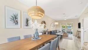 A spacious dining area with a wooden table and grey upholstered chairs, a hanging woven light fixture, and framed botanical artwork on the walls. - Coral Oasis – Spacious 3BR Retreat, Shared Pool, Dog Friendly, Parking, Minutes to Downtown Key West (Key West)