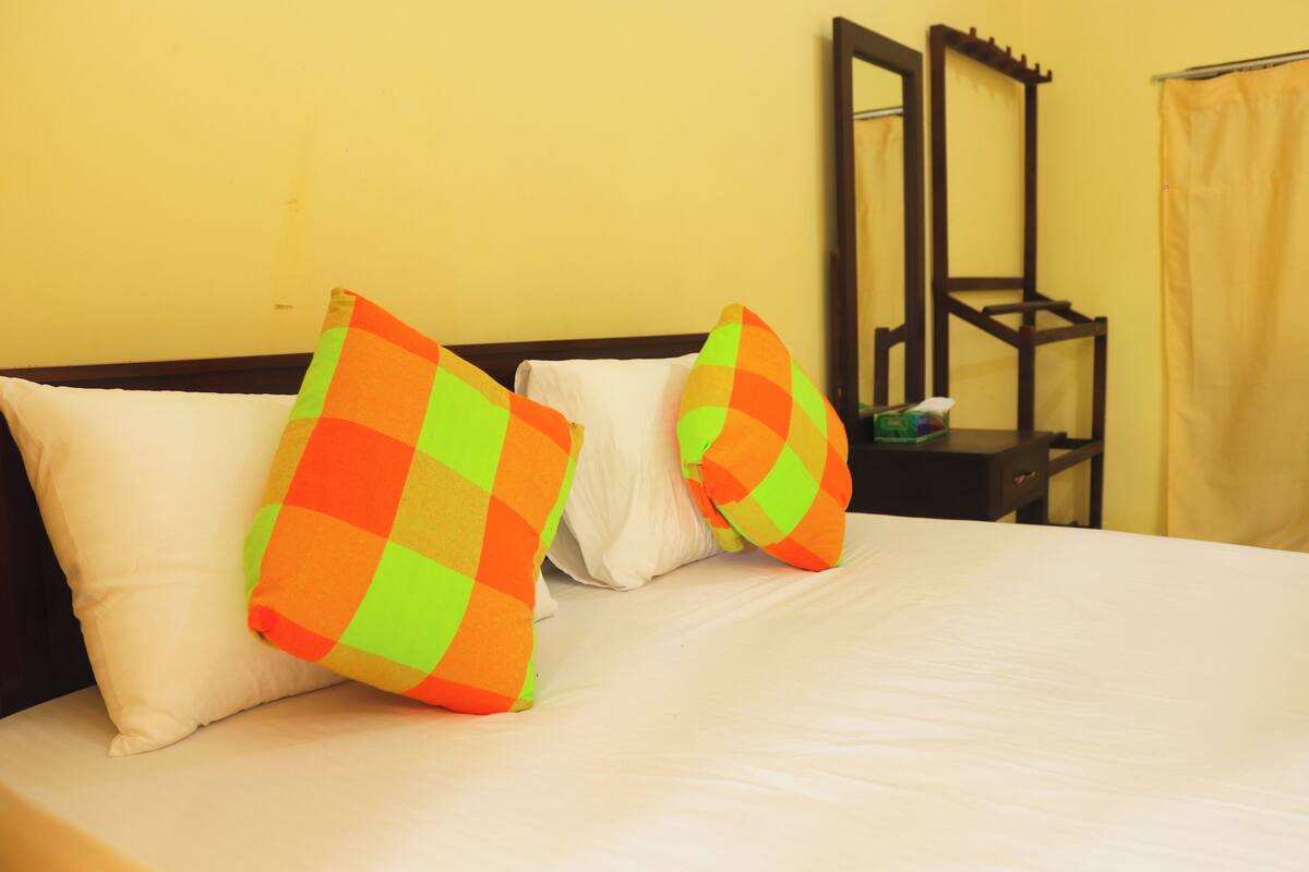 A bed with white pillows and two colorful pillows, a wooden nightstand with a mirror, and a yellow wall.