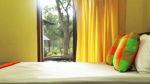 A bed with a white sheet and colorful pillows, a window with yellow curtains, and a view of trees and a building outside. - Leon Lakeside Hotel (Sella Kataragama)