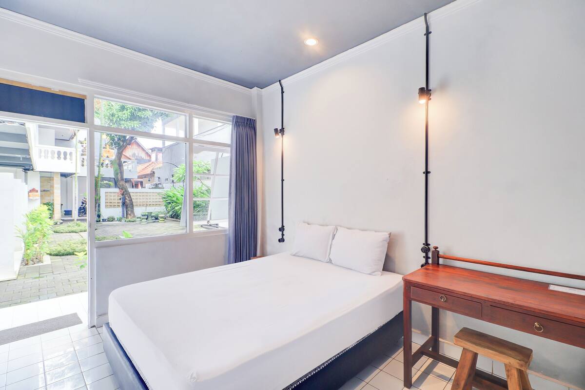 A bedroom with a bed, a wooden desk, a bench, and a window with curtains.