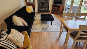 Un salon avec un canapé noir, des coussins jaunes et blancs, une table basse en bois, un téléviseur posé sur un meuble en bois, et un coin repas avec une table et des chaises en bois.