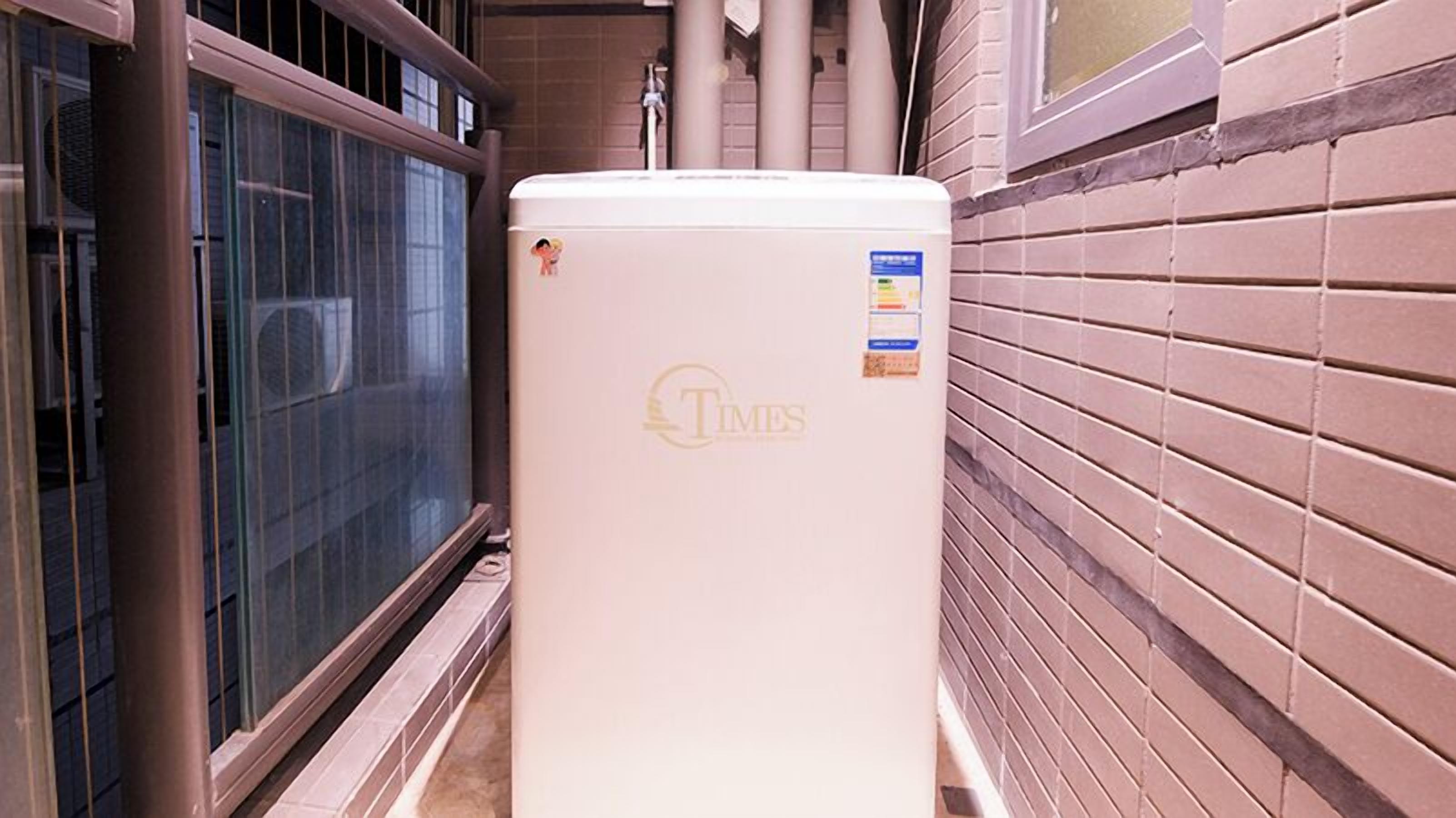A white refrigerator with a Times logo sticker, located in a narrow hallway with a window and a brick wall.