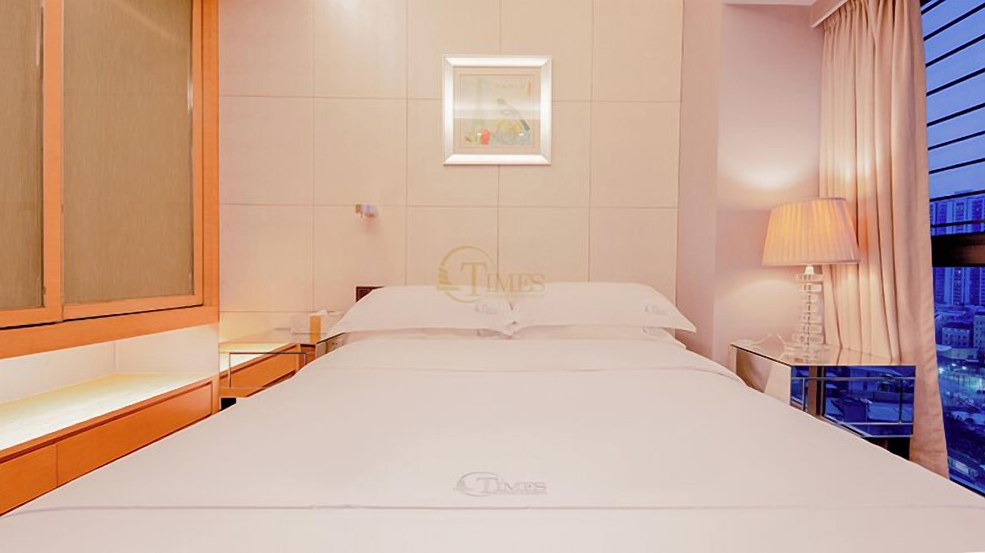 A hotel room with a bed, a nightstand, a lamp, and a window with a city view.