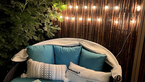 Un espacio de estar al aire libre con forma circular, con un cojín color turquesa y almohadas estampadas, rodeado de follaje y una pared de bambú con luces colgantes.