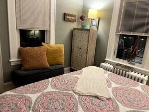 Une chambre à coucher avec un lit, une table de chevet, une lampe, un radiateur et une fenêtre avec des stores.