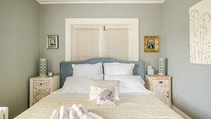 Un dormitorio con una cama, dos mesitas de noche, un ventanal con persianas y dos cuadros enmarcados en la pared.