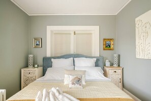 Une chambre à coucher comprenant un lit, deux tables de chevet, une fenêtre avec des volets et des œuvres d’art murales.