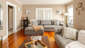 Una sala de estar con un sofá seccional gris, una mesa de centro con objetos decorativos, un sillón blanco y una ventana con persianas.