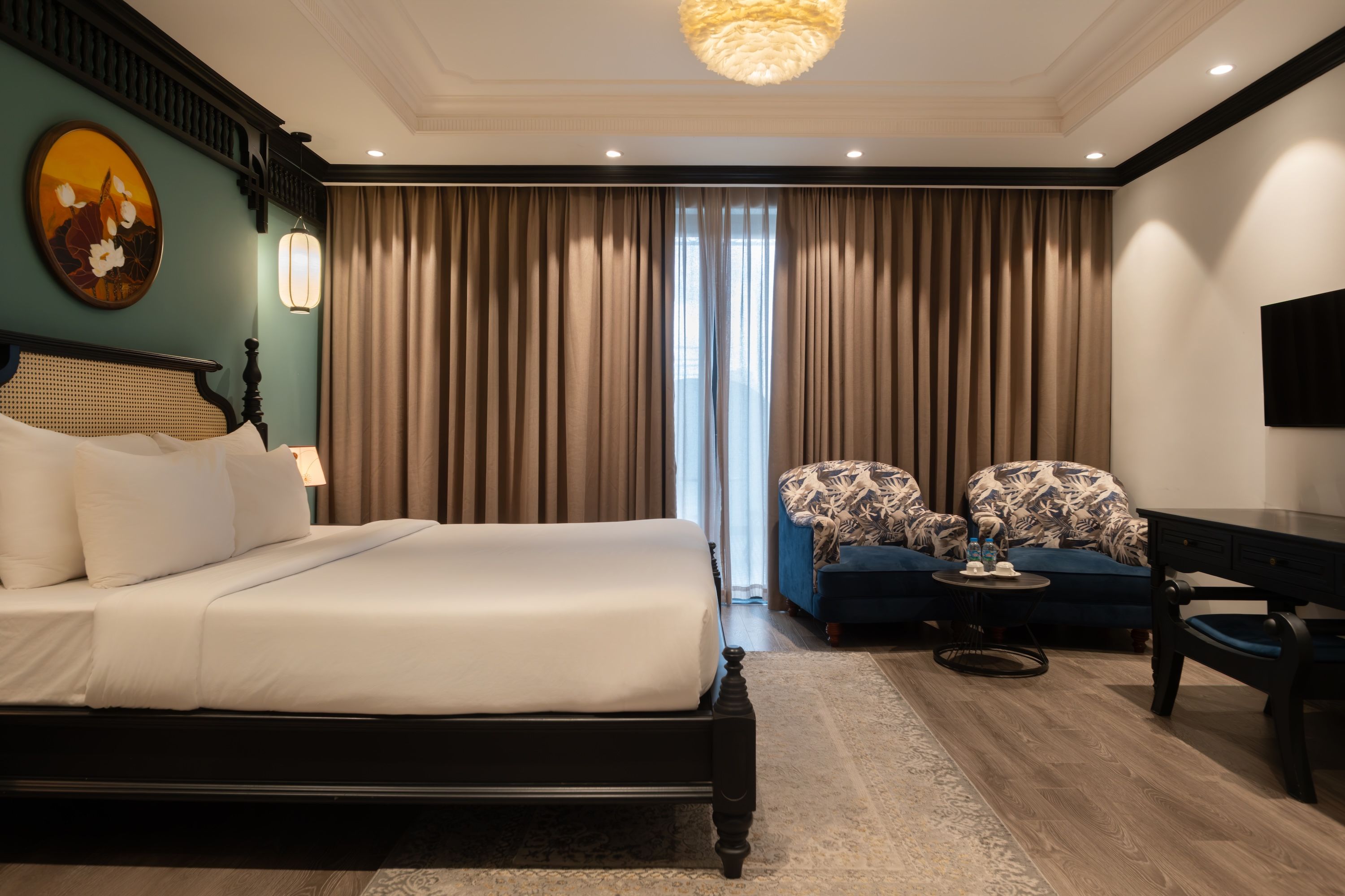 Habitación de hotel con una cama grande, dos sillones estampados, una mesita con botellas y un televisor en la pared.