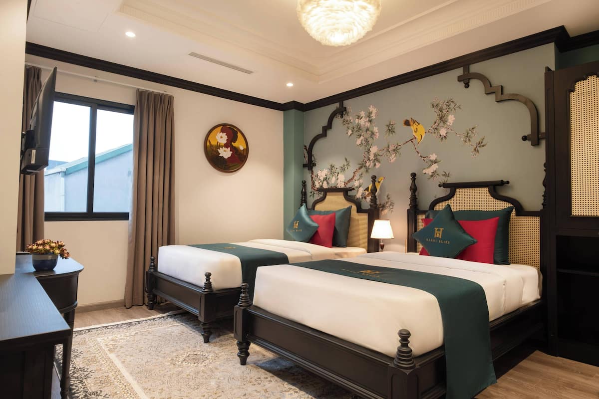 A hotel room with two beds, a large window with curtains, a decorative wall, and a small table with a vase.