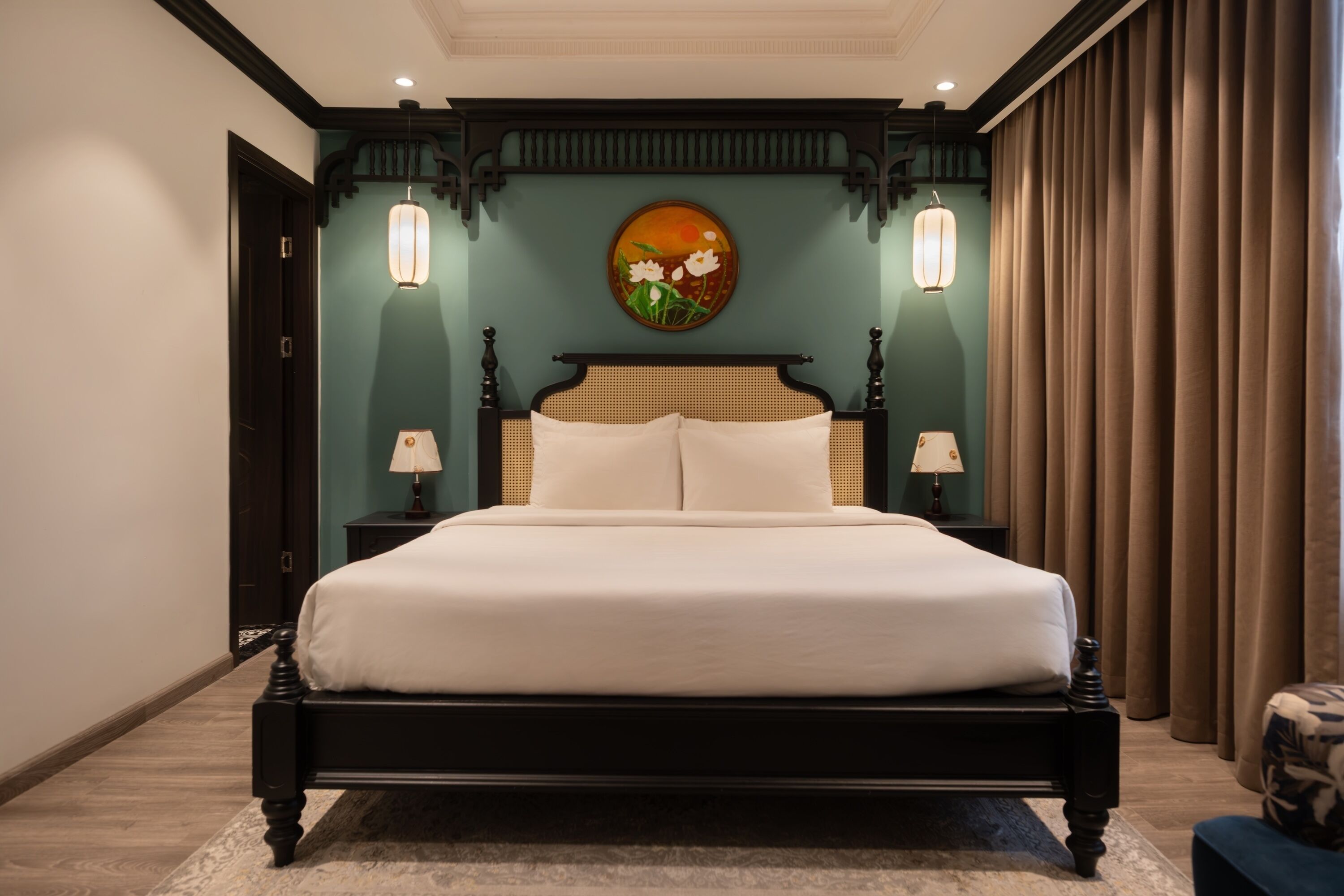 Een groot bed met een donker frame, twee bedlampen, een schilderij aan de muur en een raam met gordijnen.