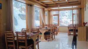 A wooden interior with multiple tables and chairs, large windows with curtains, and a view of a snowy outdoor area. - Hotel Nayra Manali (Manali)