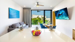 A modern living room with a sofa, a dining table with fruit, a TV, and a balcony with a view.