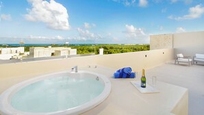 A rooftop terrace with a hot tub, lounge chairs, and a view of the ocean and greenery.