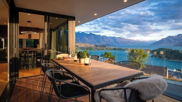 Un comedor con una mesa y sillas de madera, vista a un lago y montañas, y una pared de vidrio.