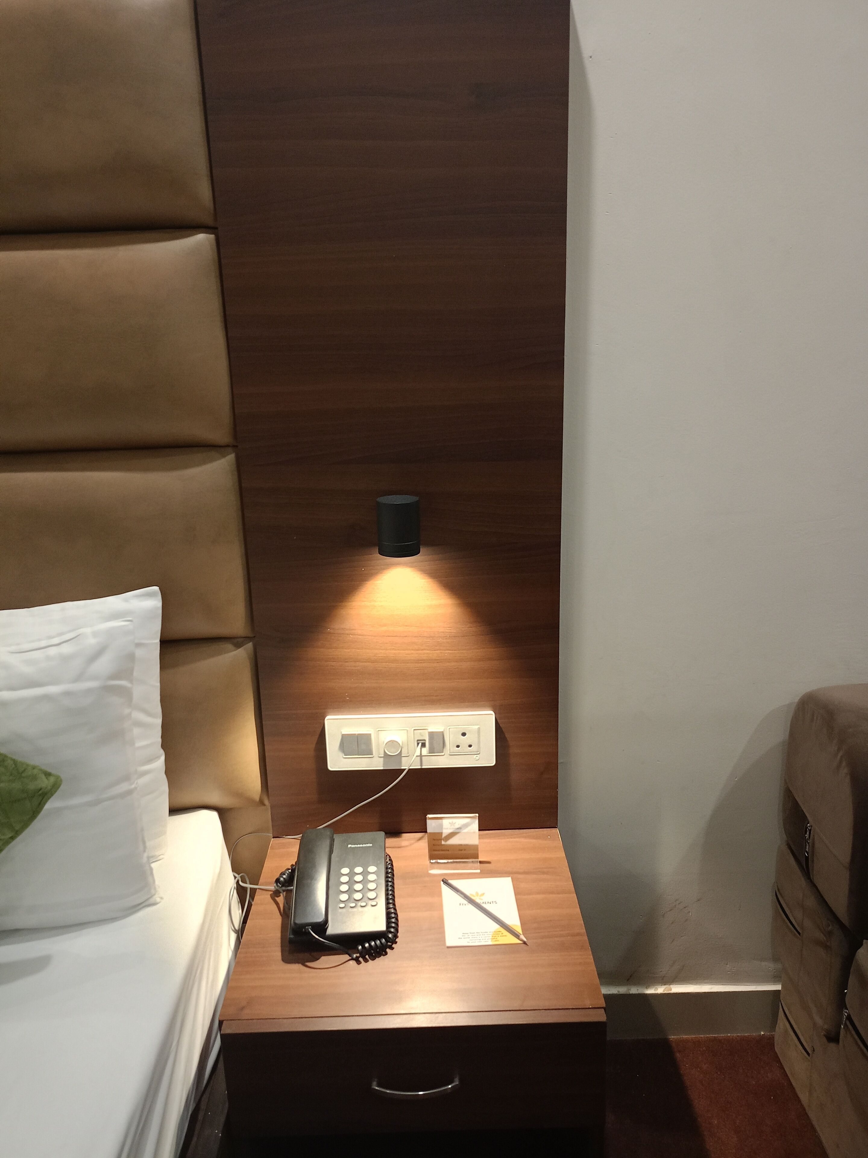 Een net opgemaakt bed met een groen kussen, een nachtkastje met een telefoon, een lamp en een stopcontact.
