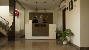 A reception desk with a marble top and a sign that reads "RECEPTION". There are two people behind the desk, a plant to the right, and a staircase to the left. - Five Elements Hotel Haridwar (Haridwar)