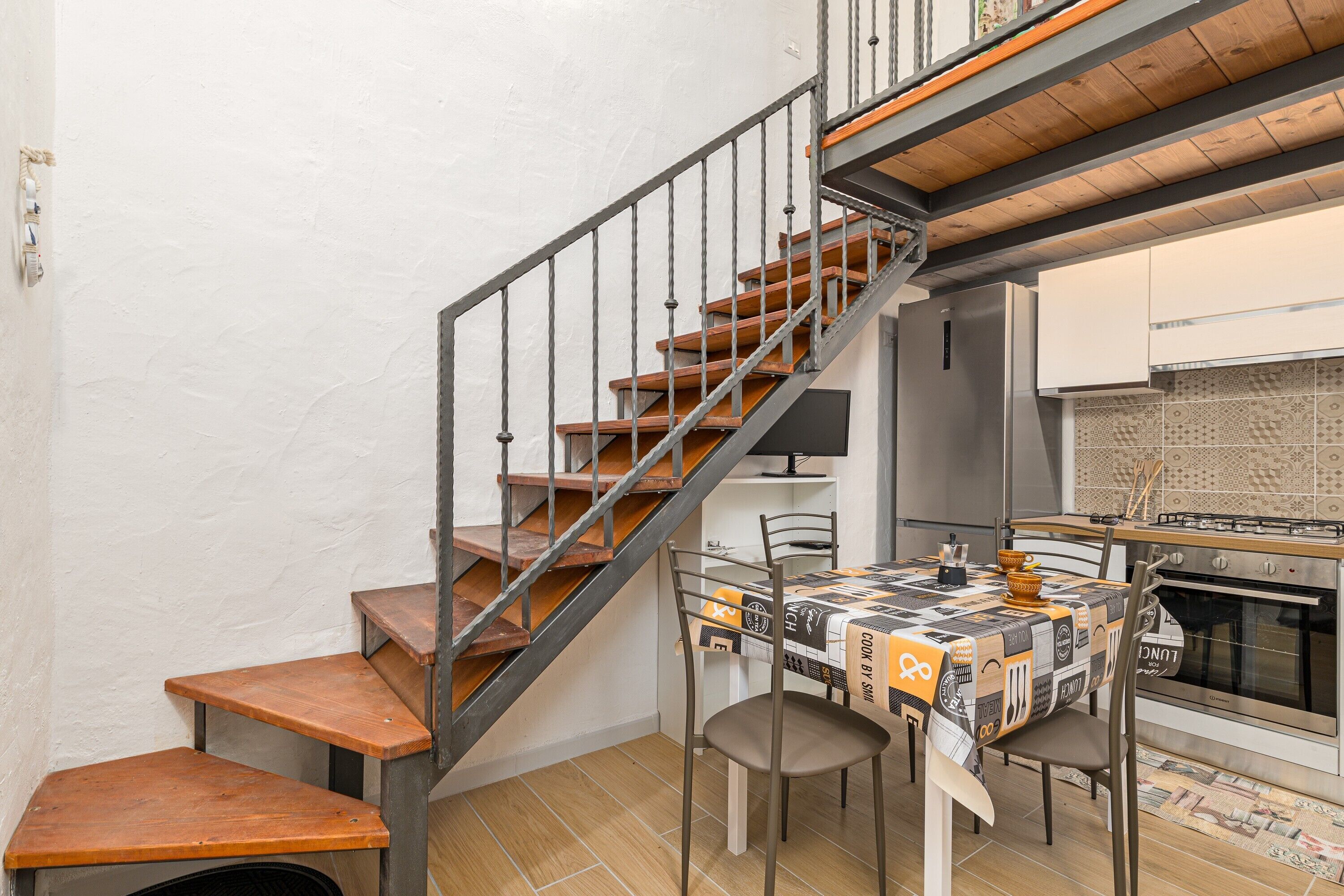A compact kitchen with a wooden staircase, a dining table with chairs, and a tiled backsplash.