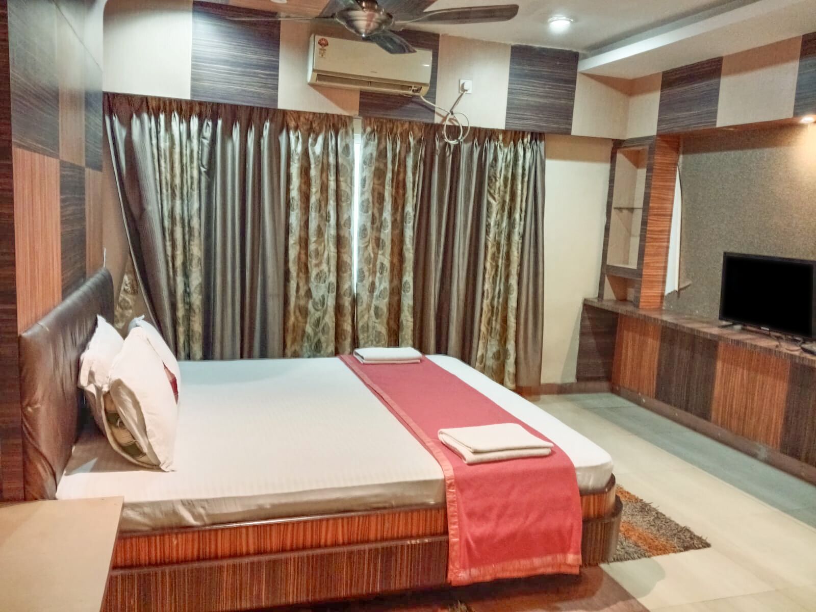 A hotel room with a bed, a television, an air conditioning unit, and a ceiling fan.