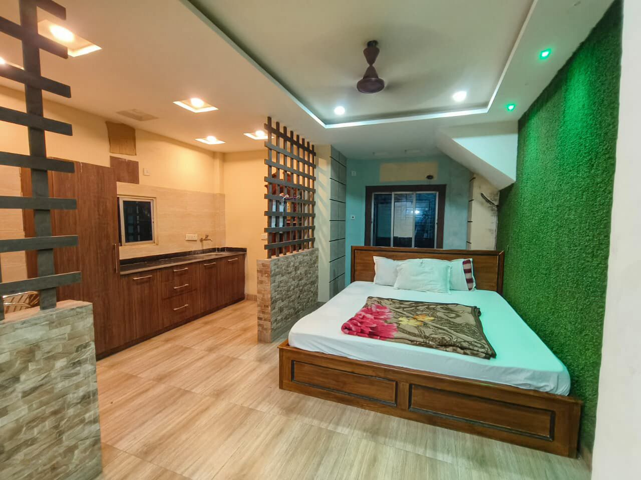 A bedroom with a bed, a wooden headboard, a green wall, a kitchen area with cabinets, and a ceiling fan.