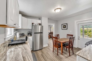 A modern kitchen with white cabinets, a stainless steel refrigerator, and a dining area with wooden chairs. - Pitcher's Paradise Apt 3 - All Star Baseball Rentals (Oneonta)