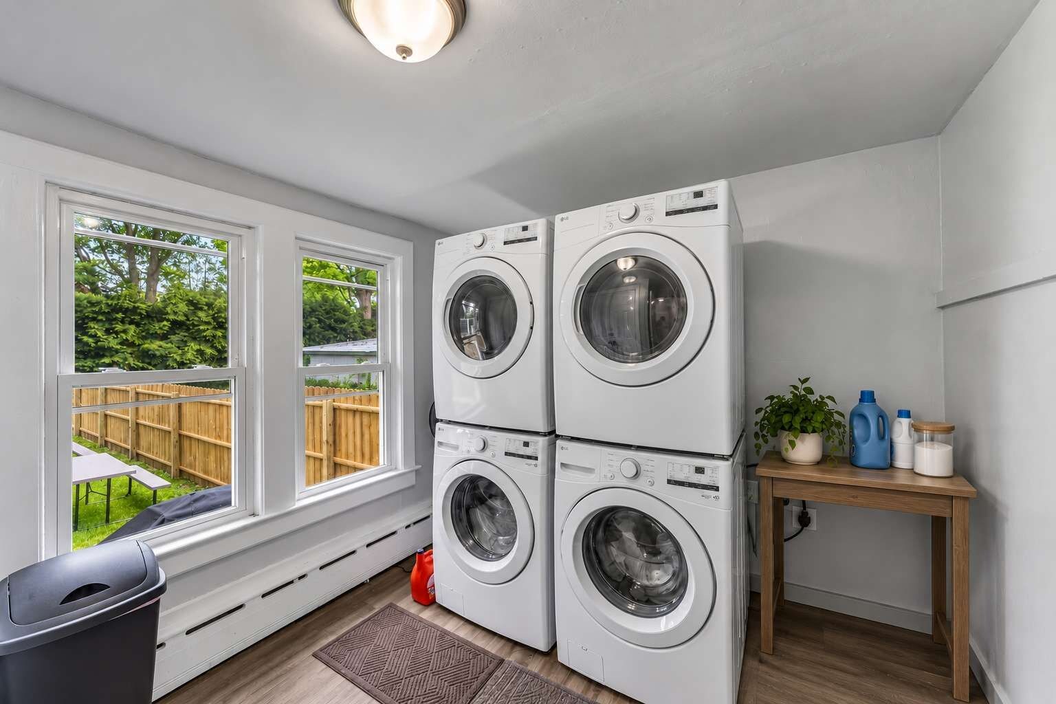 A laundry room with two stacked front-loading washing machines, a wooden table with cleaning supplies, and a window with a view of a backyard.