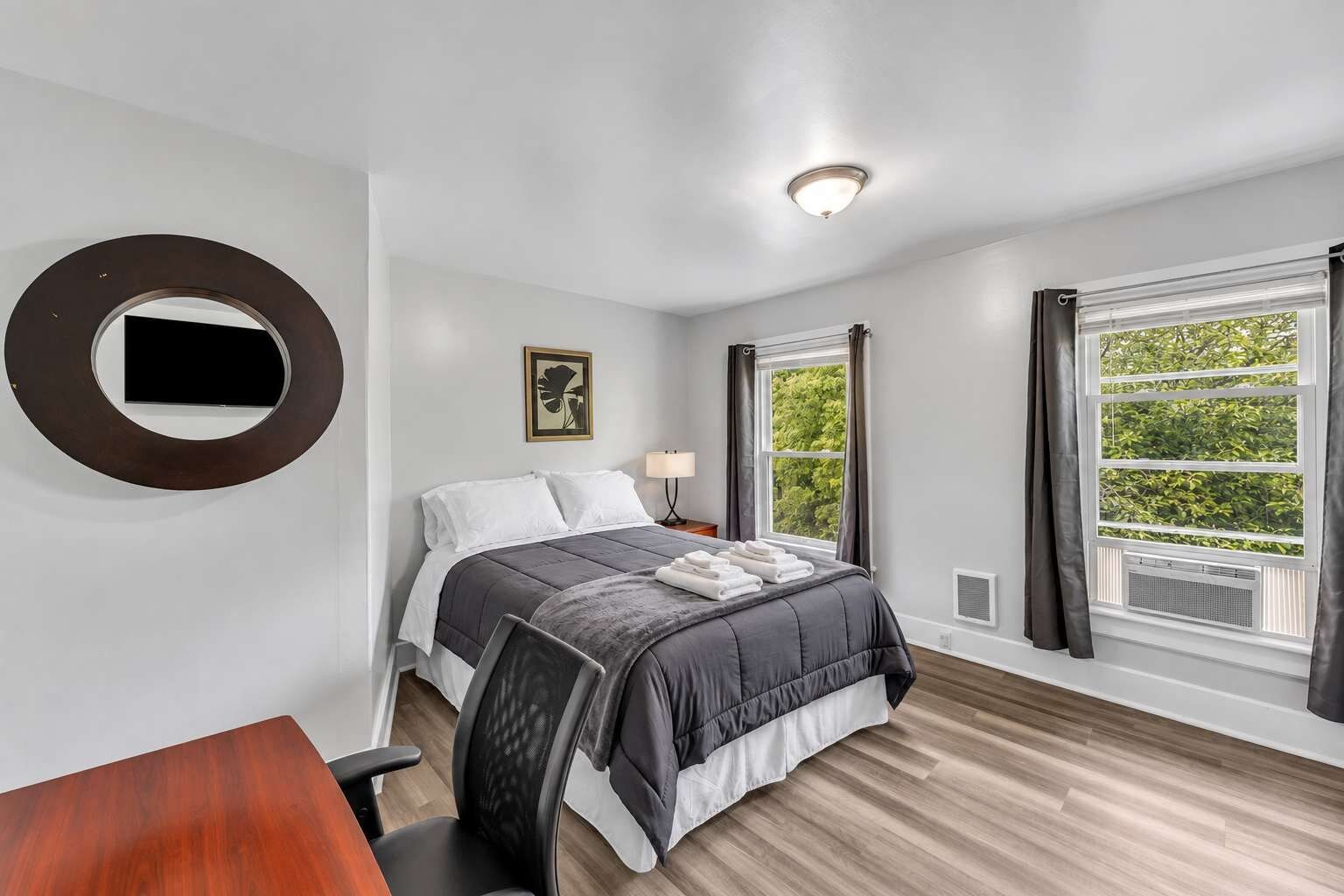 A hotel room with a bed, a desk, a chair, a round mirror, a picture on the wall, and a window with curtains.