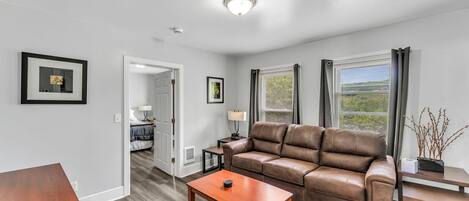 A modern living room with a leather sofa, a wooden coffee table, and a dining table. There is a door leading to a bedroom, a window with curtains, and a ceiling light.