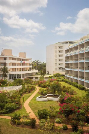 Un hotel con varios pisos, un jardín con diversas plantas y flores, y un cielo despejado.