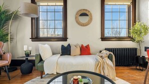 A living room with a sofa, a round mirror, two windows, a radiator, and a glass coffee table. - The Accountant | Sleeps 2 Guests | DT Drumheller | (Drumheller)