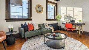 A modern living room with a green sofa, a round coffee table, a red chair, a black desk, and a large window. - The Accountant | Sleeps 2 Guests | DT Drumheller | (Drumheller)