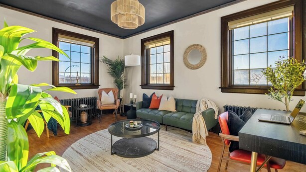 A living room with a round coffee table, a sofa, a dining table with a laptop, large windows, and a hanging light fixture. - The Accountant | Sleeps 2 Guests | DT Drumheller | (Drumheller)