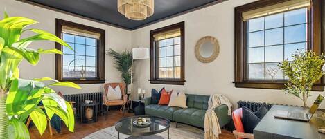 A living room with a round coffee table, a sofa, a dining table with a laptop, large windows, and a hanging light fixture.