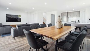 A modern living room with a wooden dining table, black upholstered chairs, a grey sofa, and a large flat-screen TV mounted on the wall. - The Pit Stop Tailem Bend (Tailem Bend)