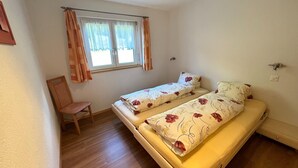 A small room with two single beds, a wooden chair, a window with curtains, and a wall socket. - Mountain Stay With Balcony (Saas Grund)